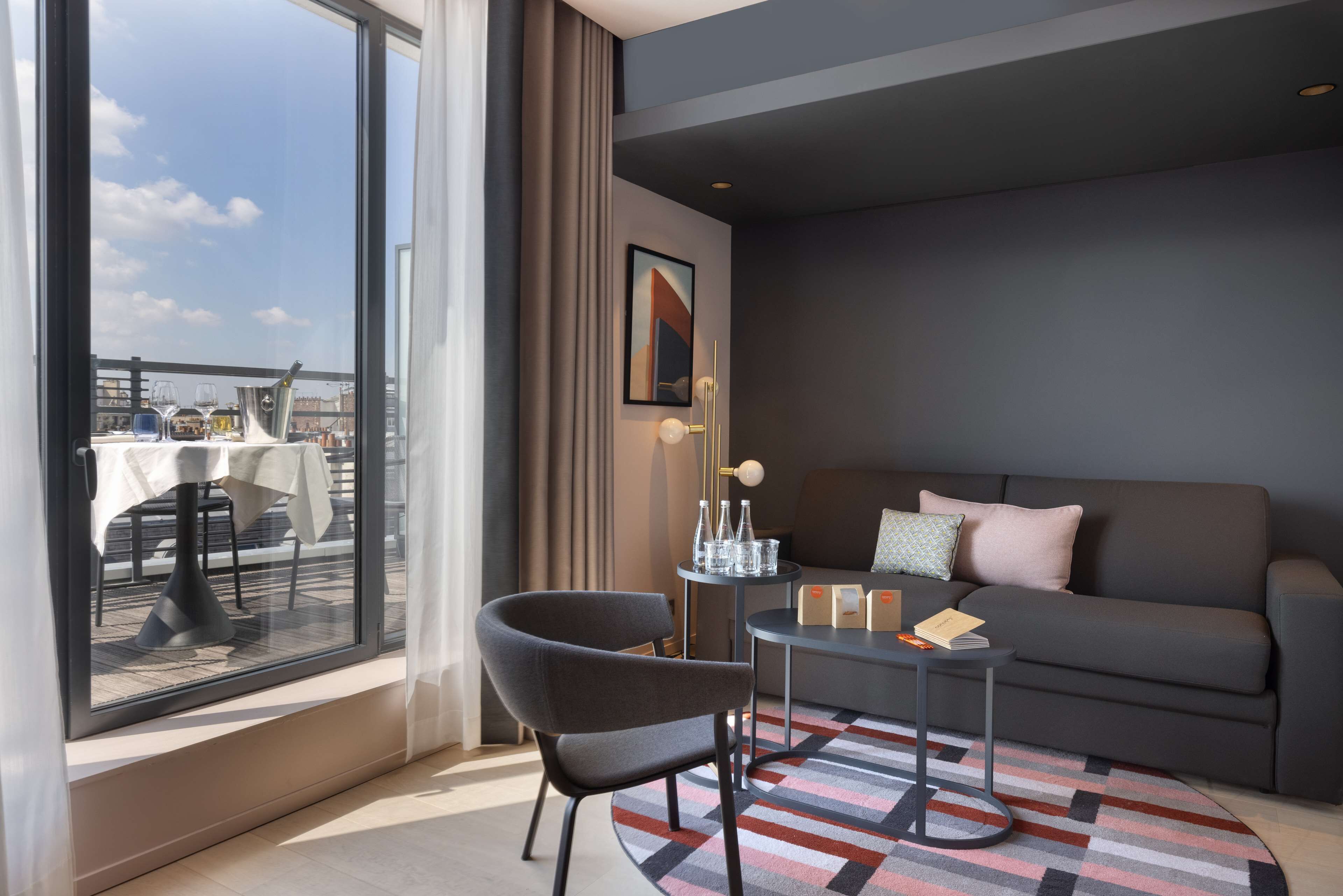 A modern hotel room with a large window, a sofa, a chair, a small table, and a patterned rug.
