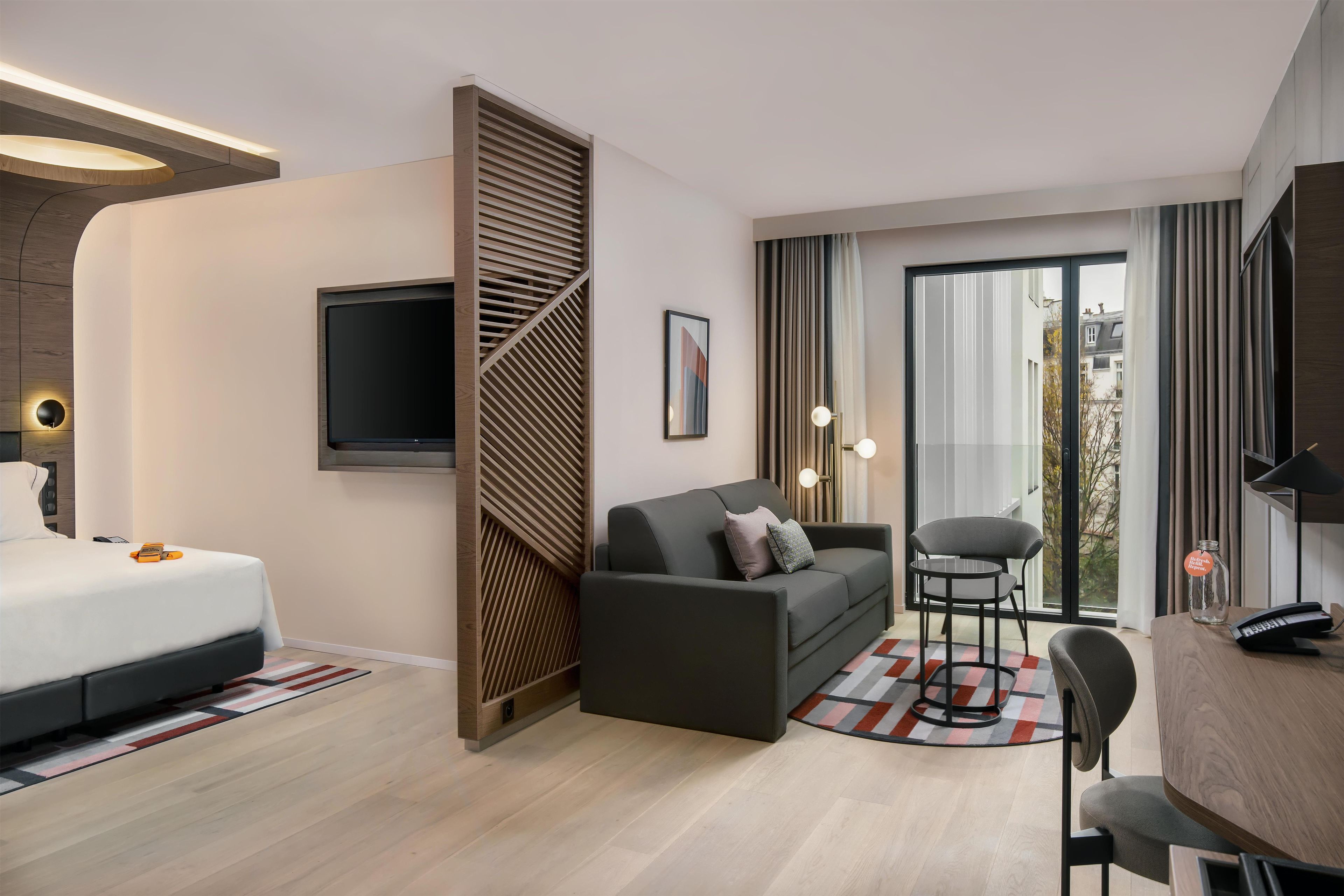 A modern hotel room with a bed, a sofa, a TV, a desk with a chair, and a view through a large window.