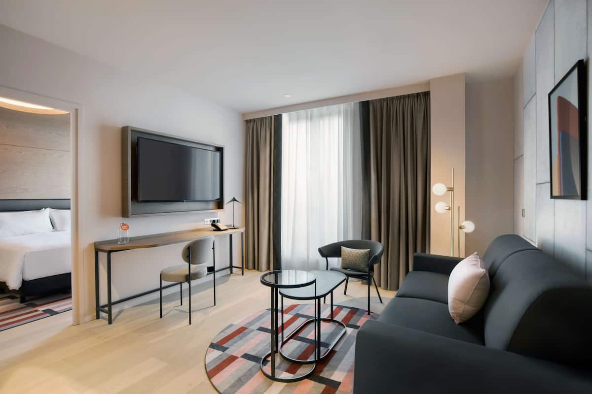 A modern hotel room with a bed, a TV mounted on the wall, a small dining area with a table and chairs, a sofa, and a large window with curtains.