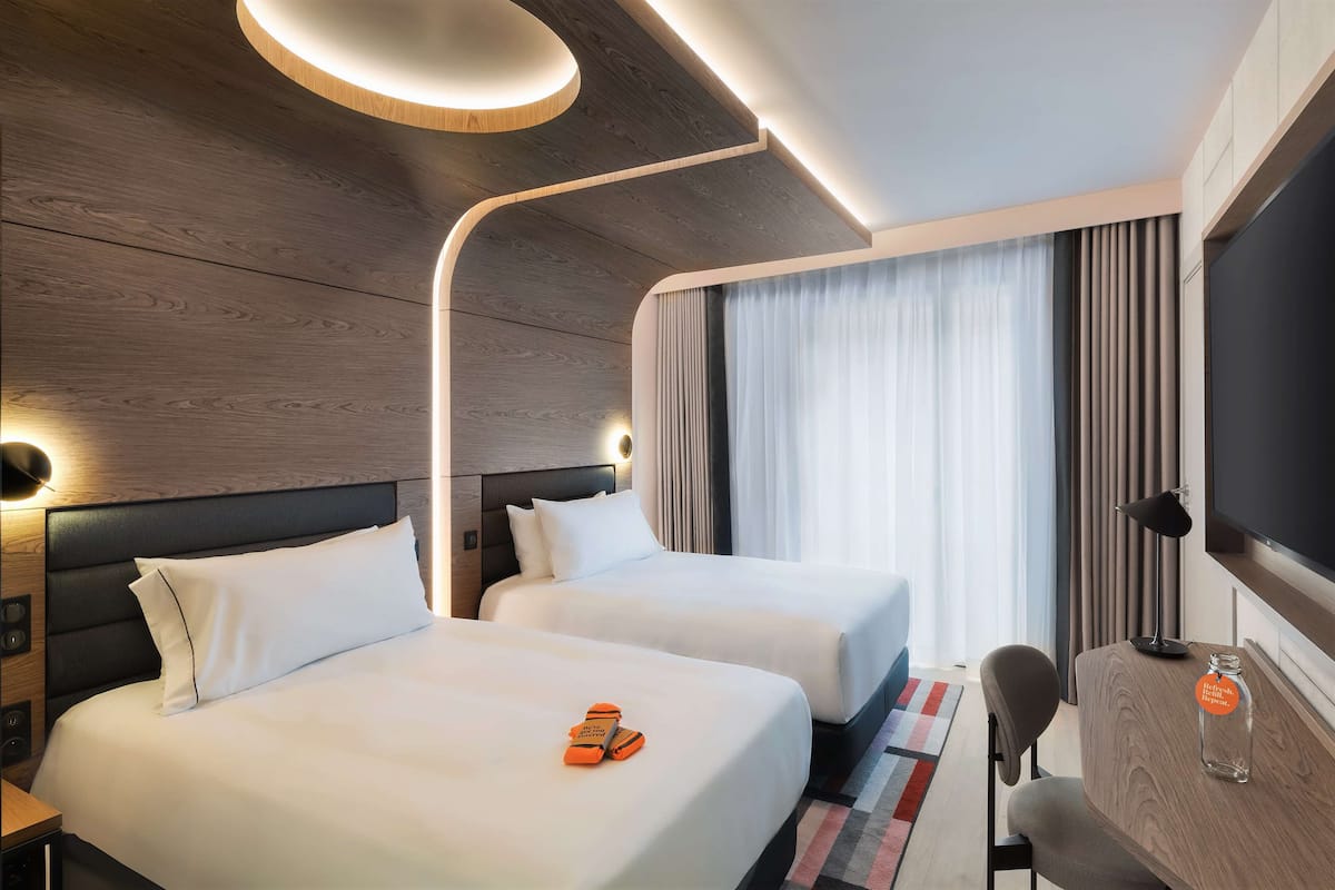 A modern hotel room with two beds, a desk, a TV, and a large window with curtains.