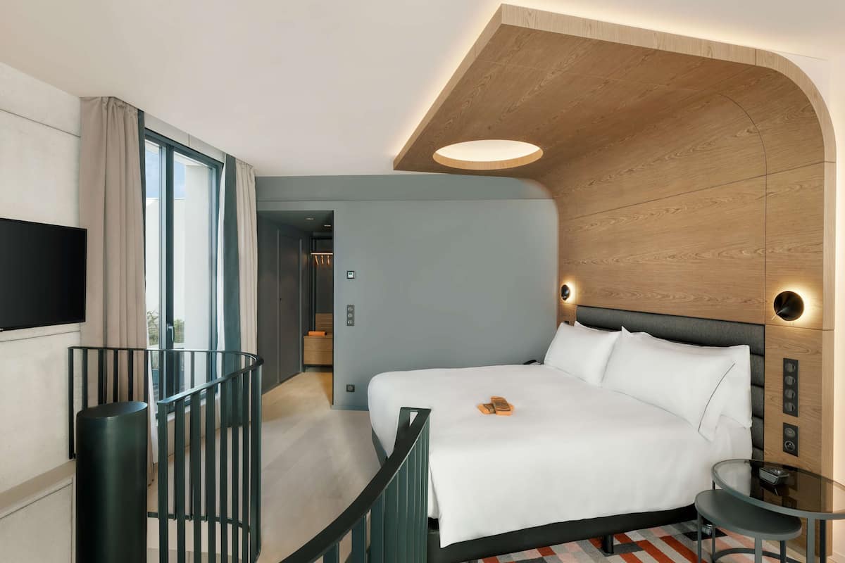 A modern hotel room with a large bed, a TV, a glass table, and a balcony with a railing.