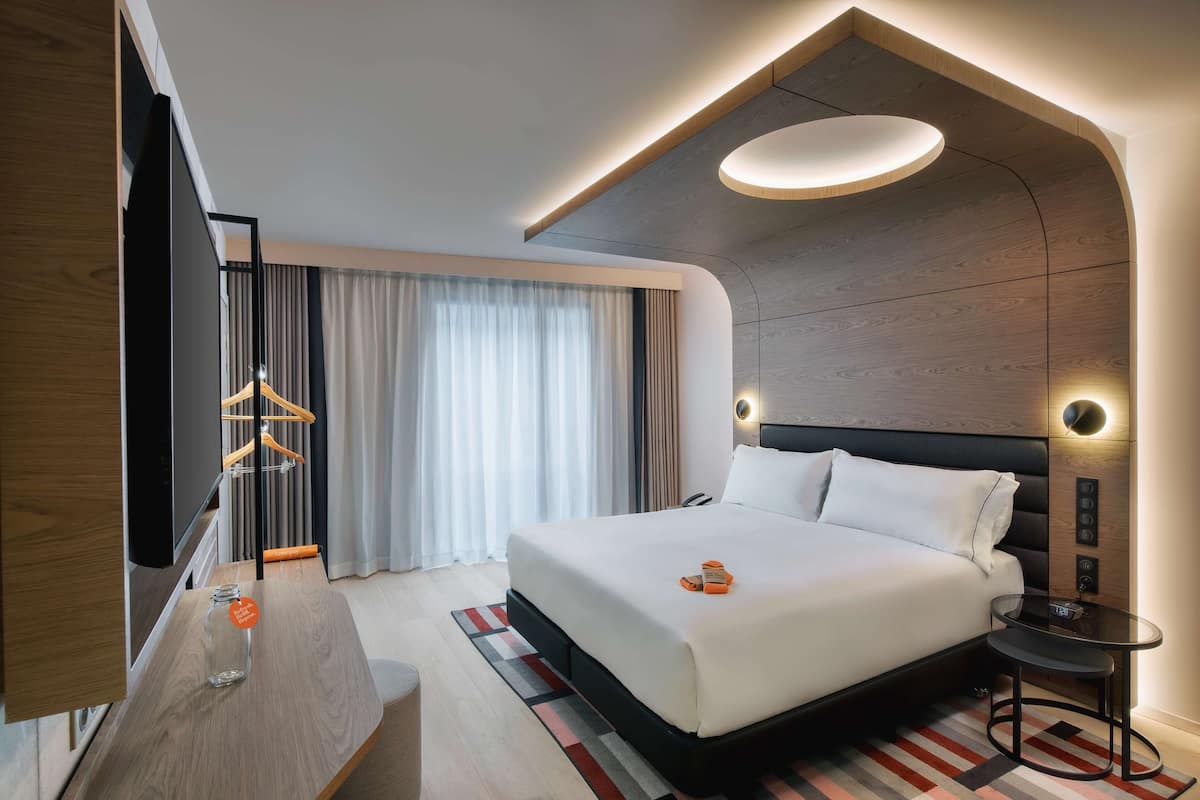 A modern hotel room with a large bed, a flat-screen TV, a desk with a glass jar, and a unique ceiling design.