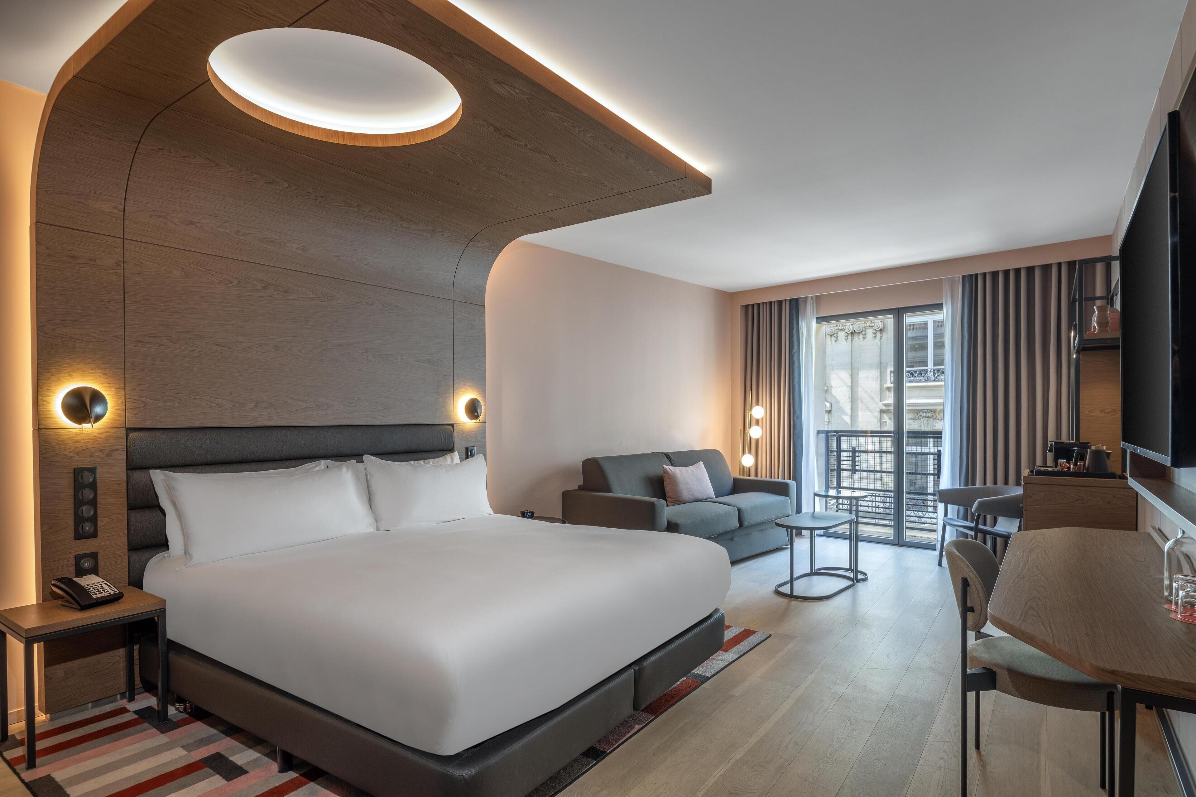 A modern hotel room with a large bed, a sofa, a dining table, and a TV.
