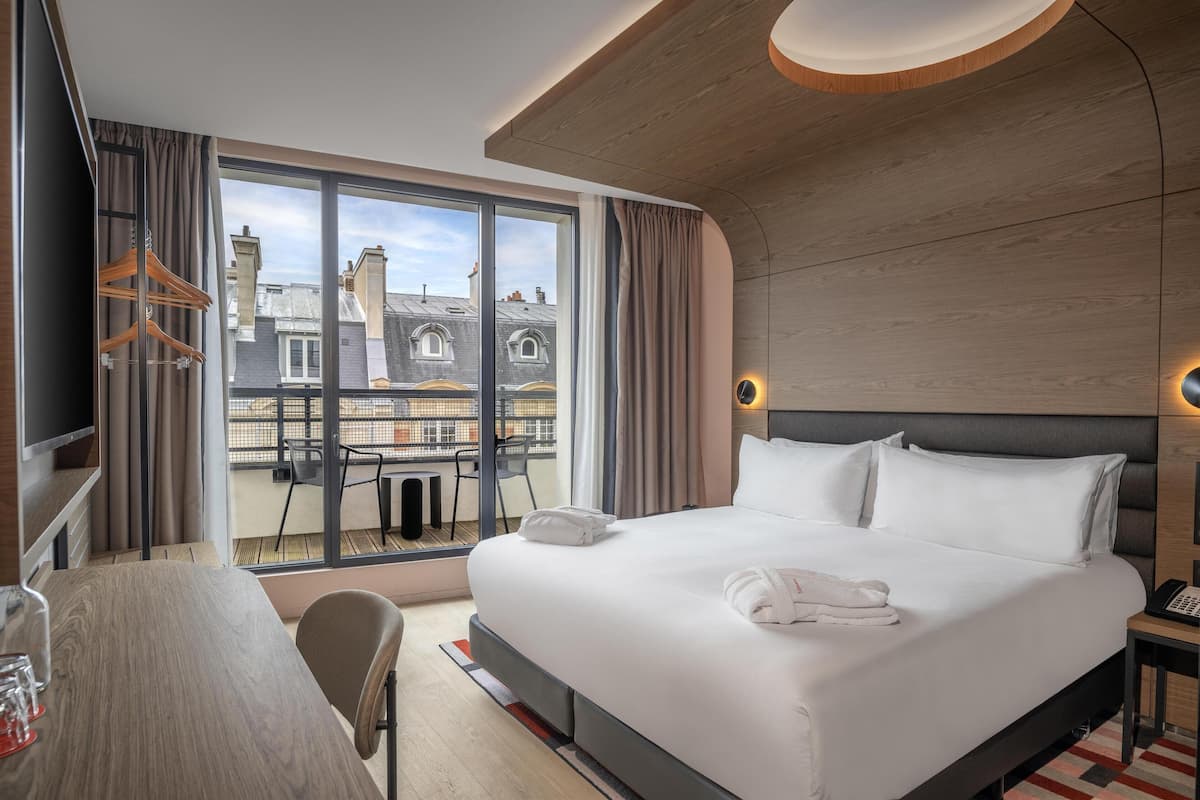 A modern hotel room with a large bed, a desk, and a view of the cityscape through floor-to-ceiling windows.