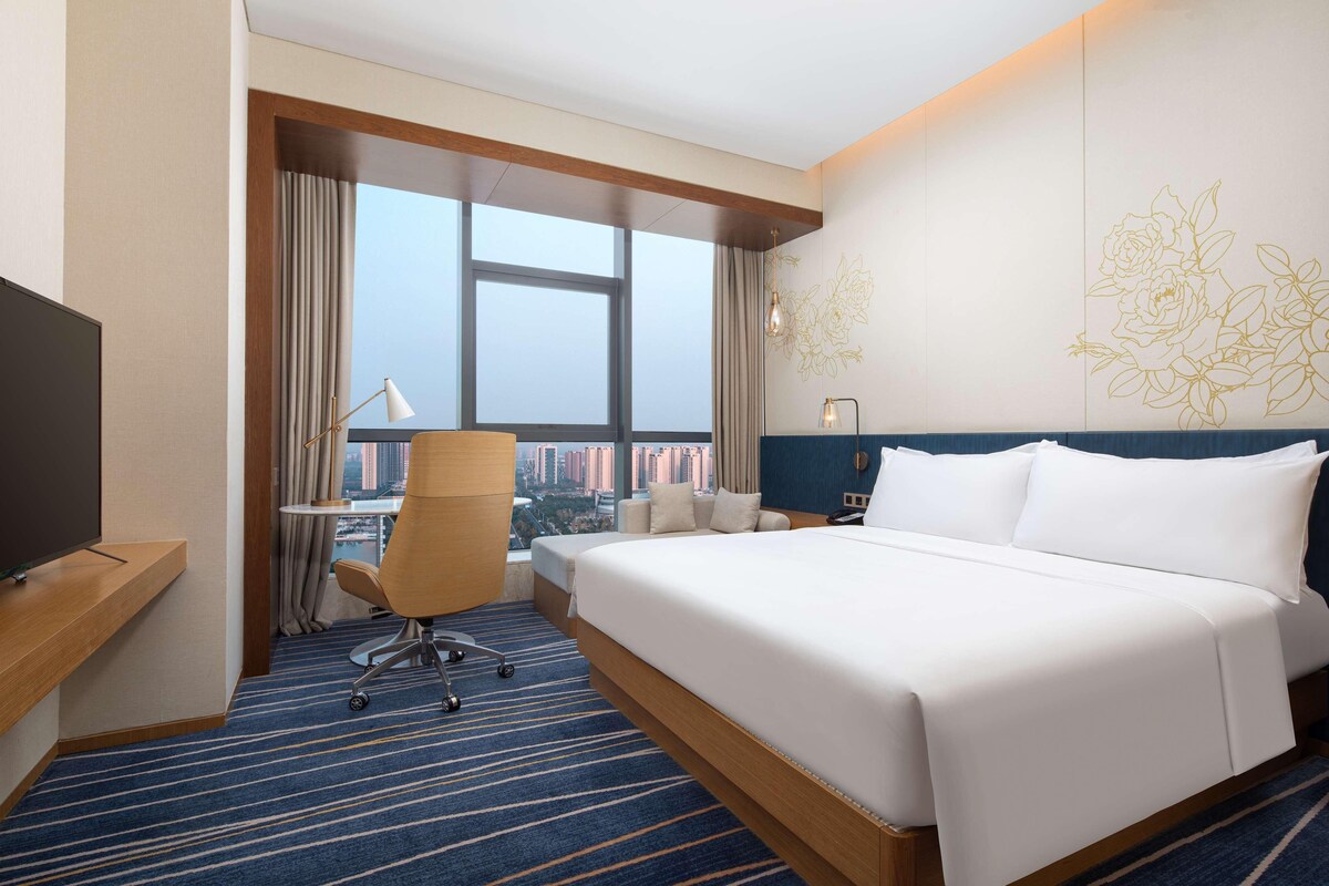 A hotel room with a large bed, a desk with a chair, a TV, and a view of the city through large windows.