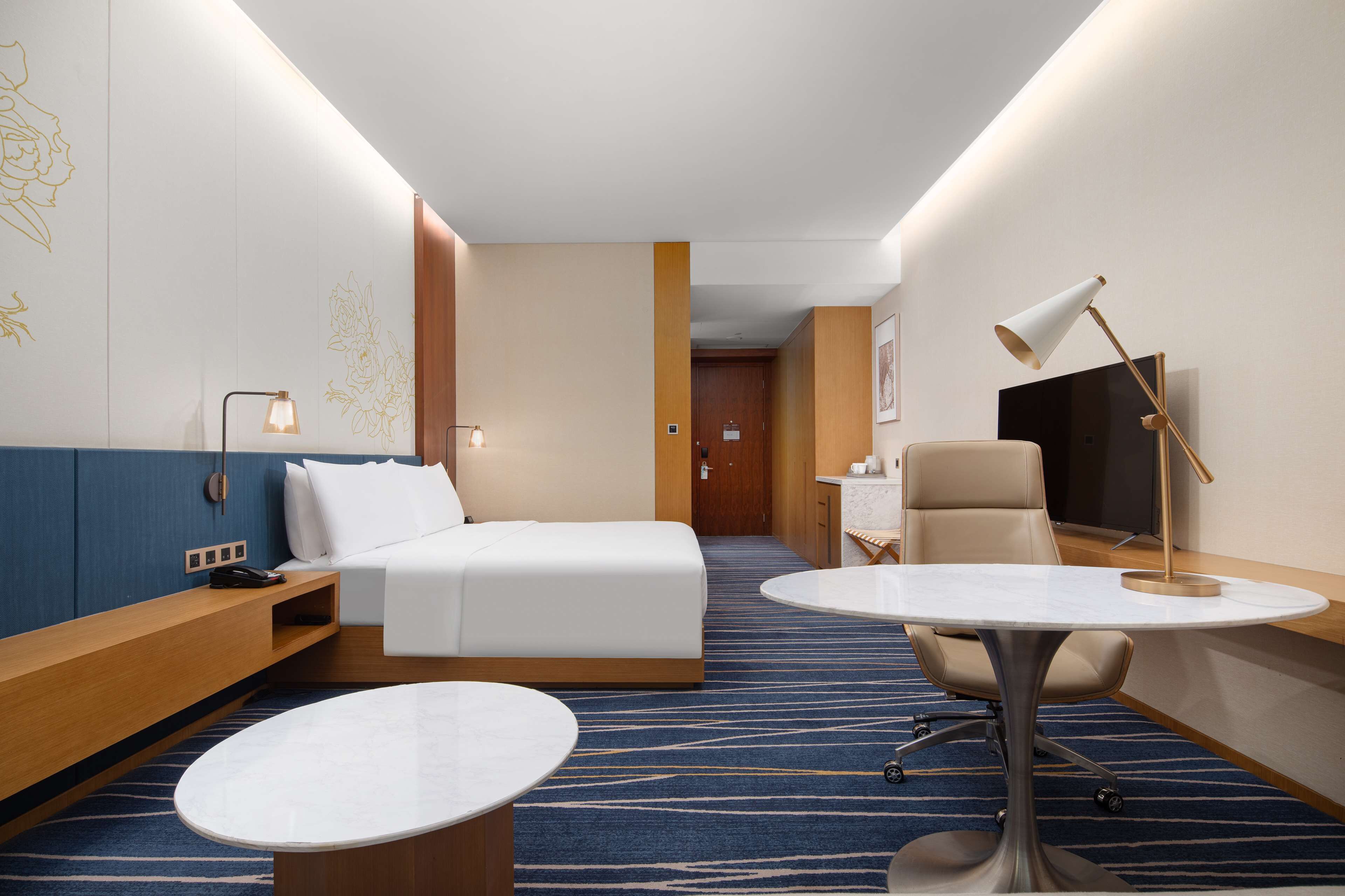 A hotel room with a bed, a desk, a chair, a round table, a TV, and a lamp.
