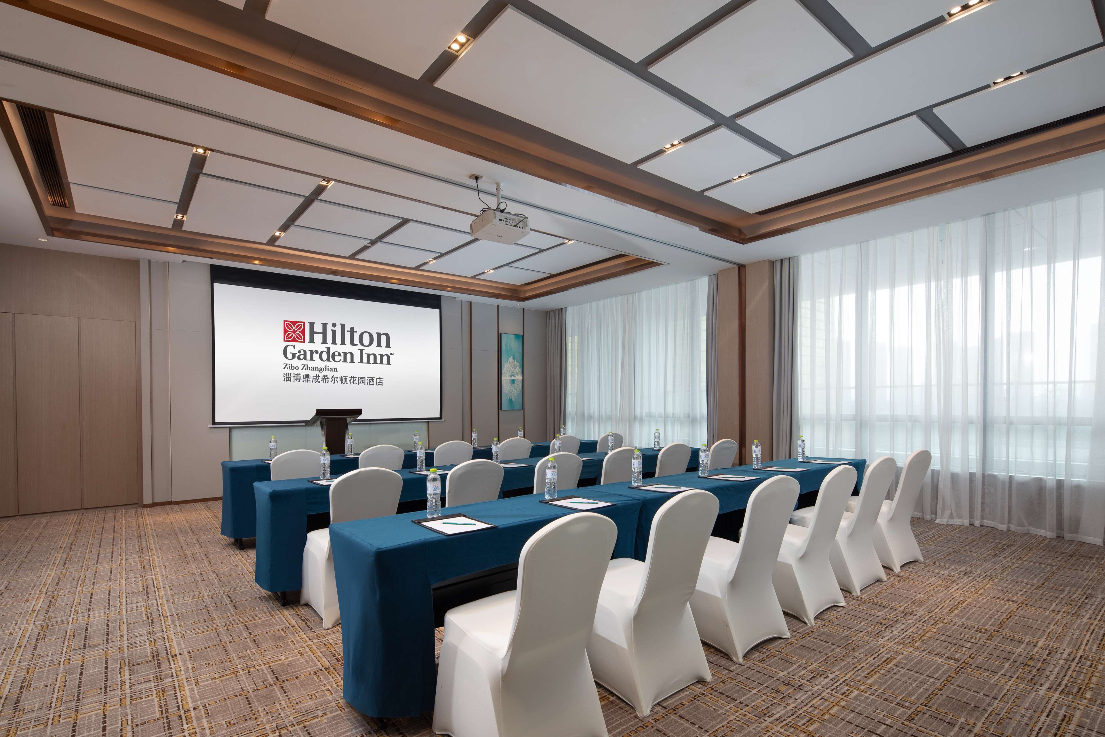 A conference room with a large screen displaying 'Hilton Garden Inn' branding, a long table with chairs, and a carpeted floor.