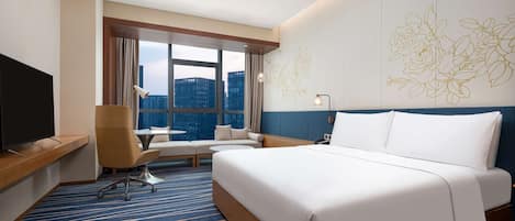 A hotel room with a large bed, a desk with a TV, a chair, a sofa, and a large window with a city view.