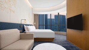 A modern hotel room with a large bed, a sofa, a flat-screen TV, and a view of tall buildings. - Hilton Garden Inn Zibo Zhangdian (Zibo)