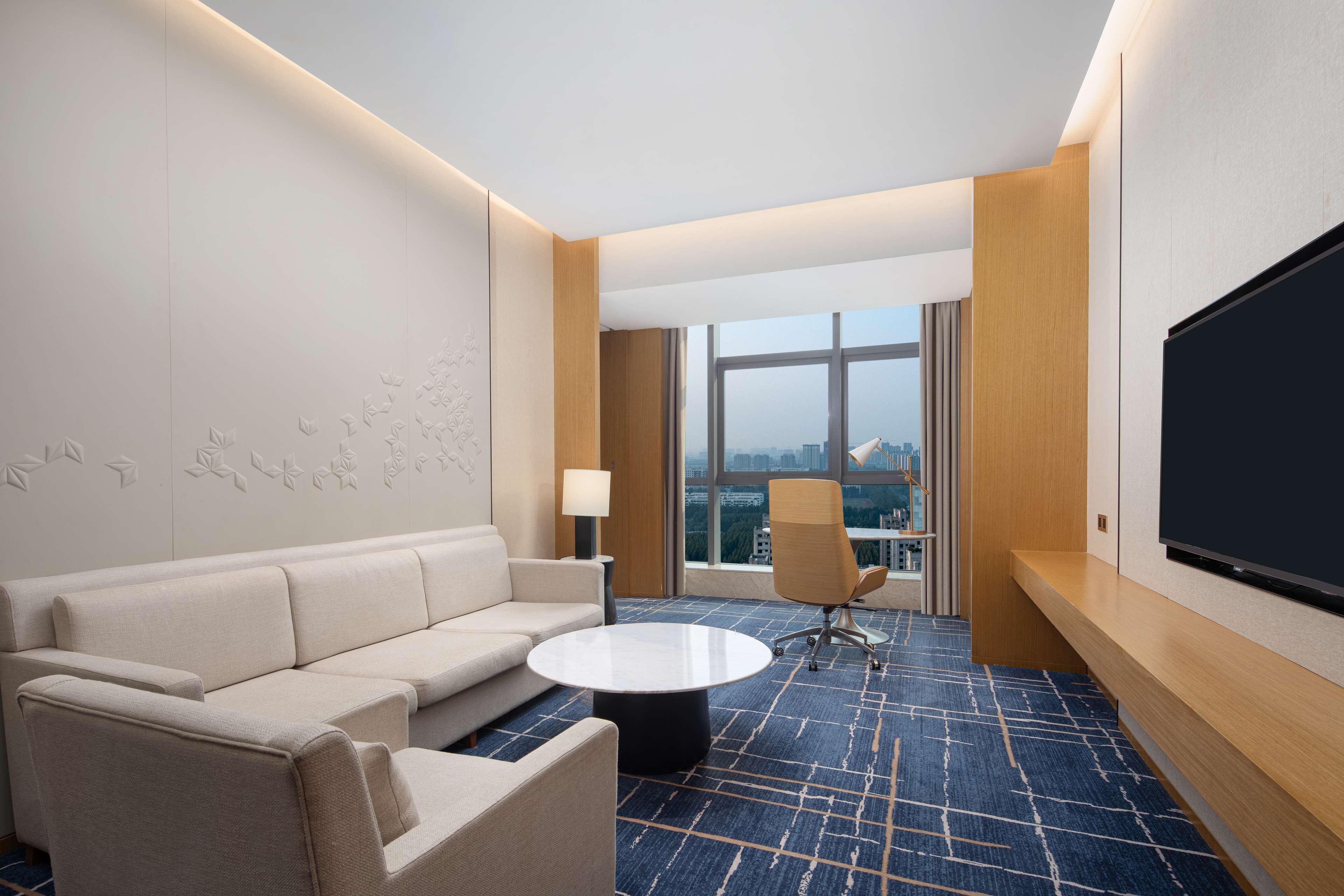 A hotel room with a sofa, a coffee table, a TV, and a large window offering a city view.