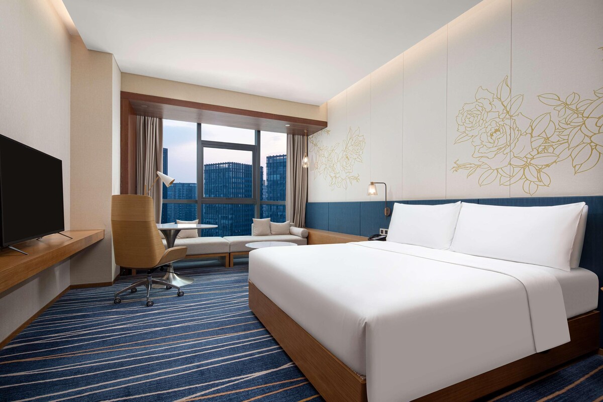 A hotel room with a large bed, a desk with a TV, a chair, a sofa, and a large window with a city view.