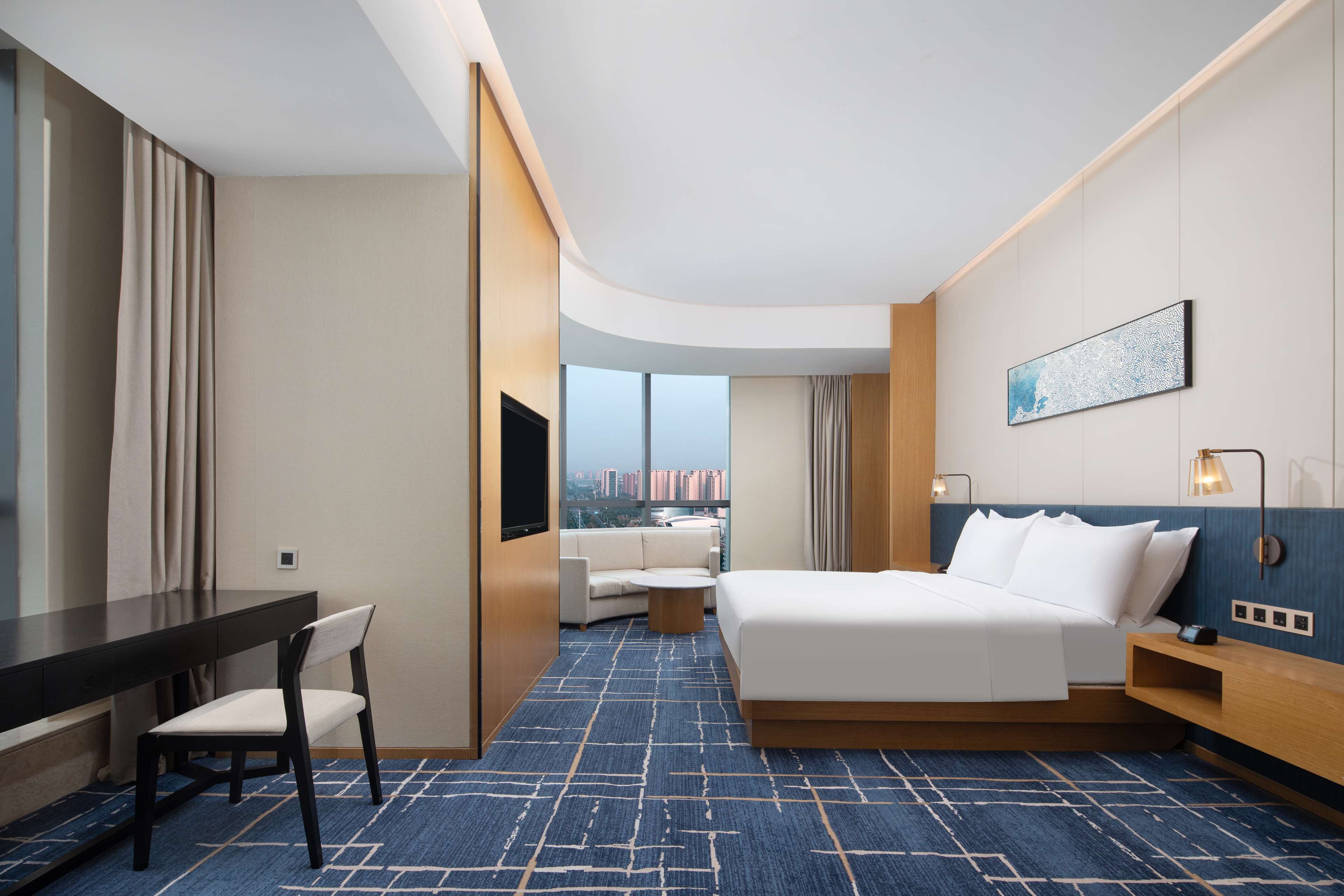 A hotel room with a bed, a desk, a chair, a small table, a TV, and a large window offering a city view.