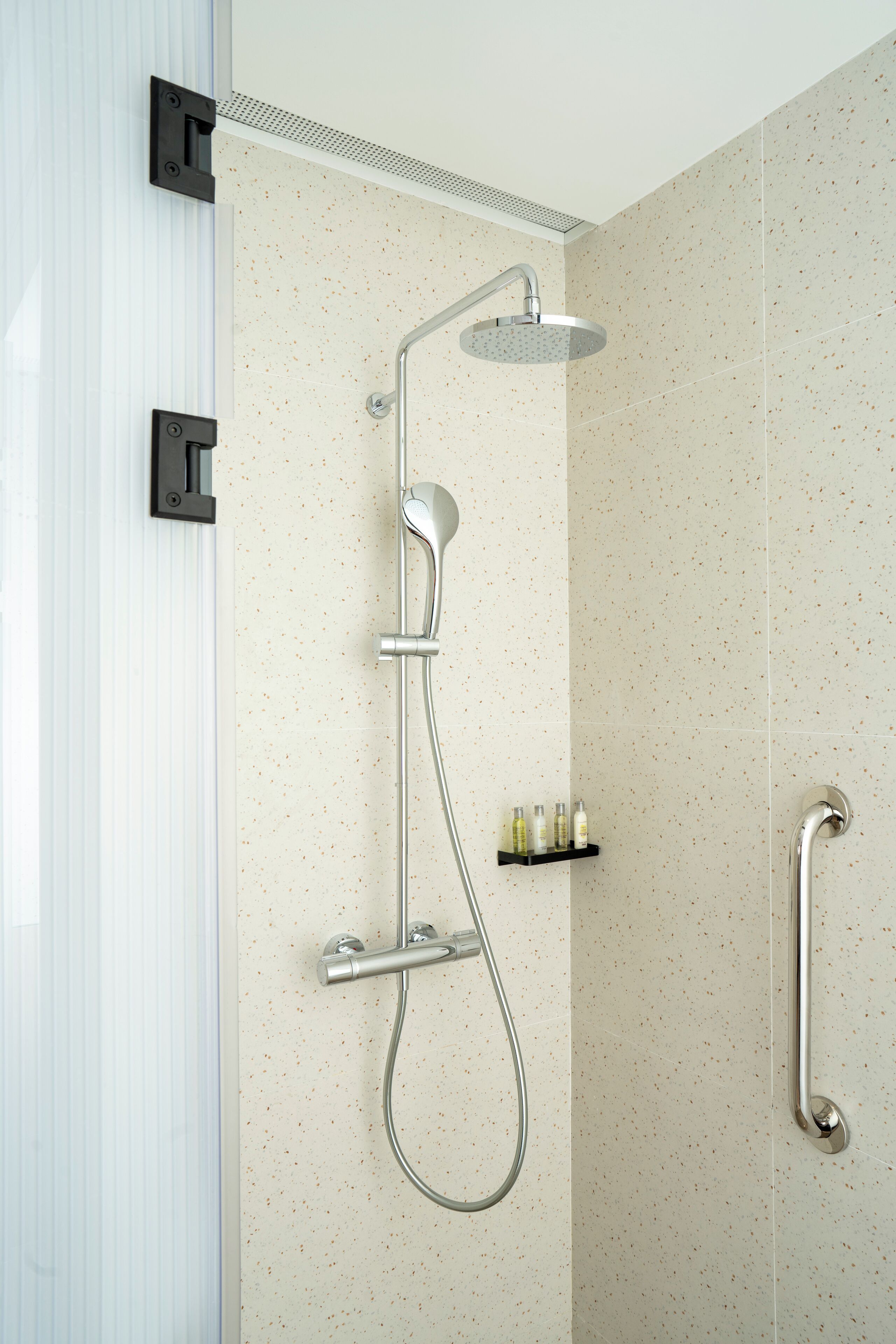 A shower with a handheld showerhead and a wall-mounted faucet, a grab bar, and a shelf with toiletries.