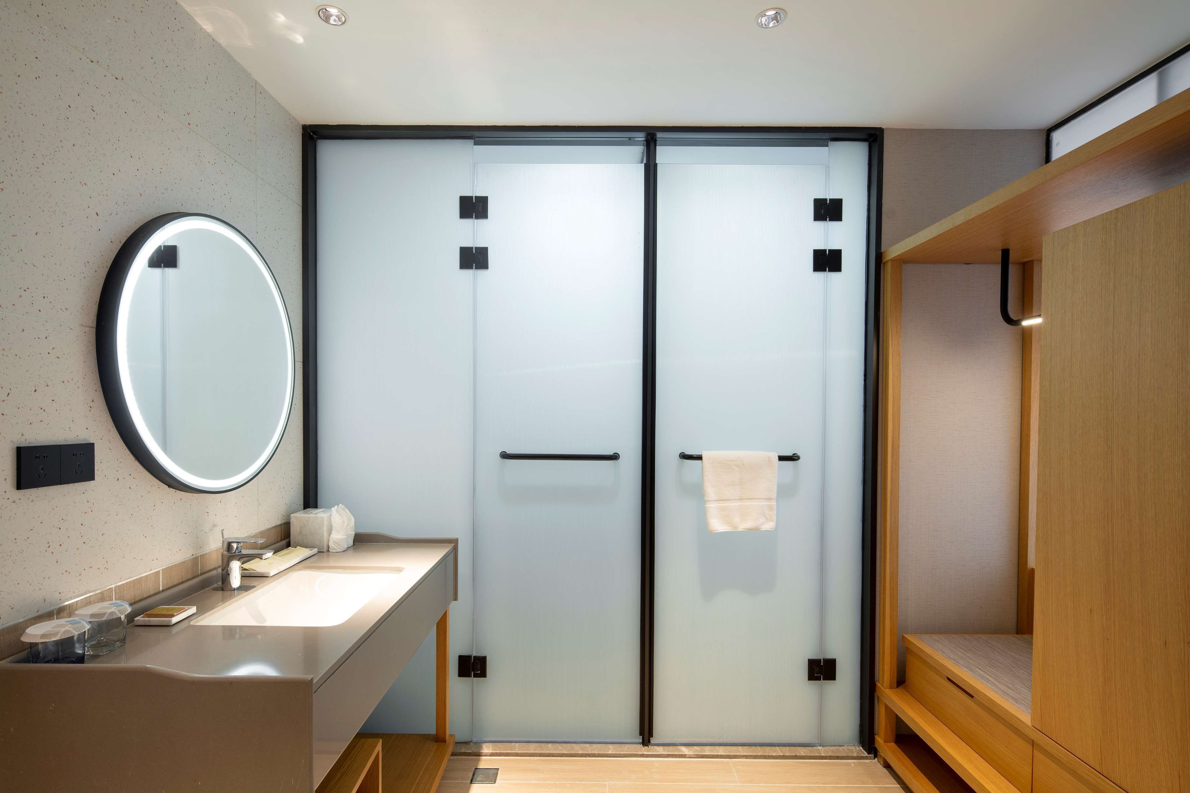 A modern bathroom with a glass door, a round mirror, a wooden bench, and a sink with a towel and soap dispenser.