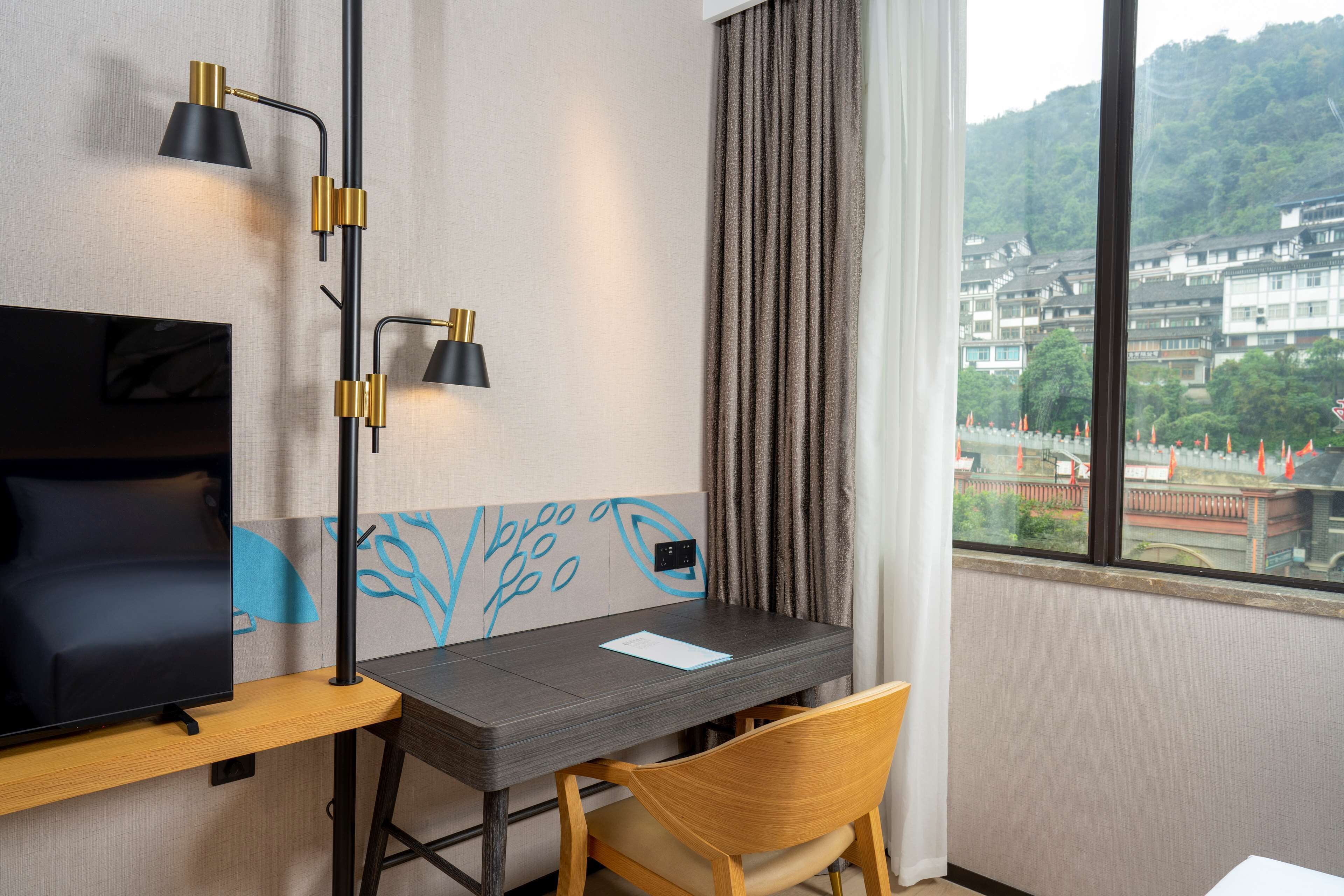 A hotel room with a TV on a wooden stand, a desk with a chair, a black lamp, and a window with a view of buildings and trees.