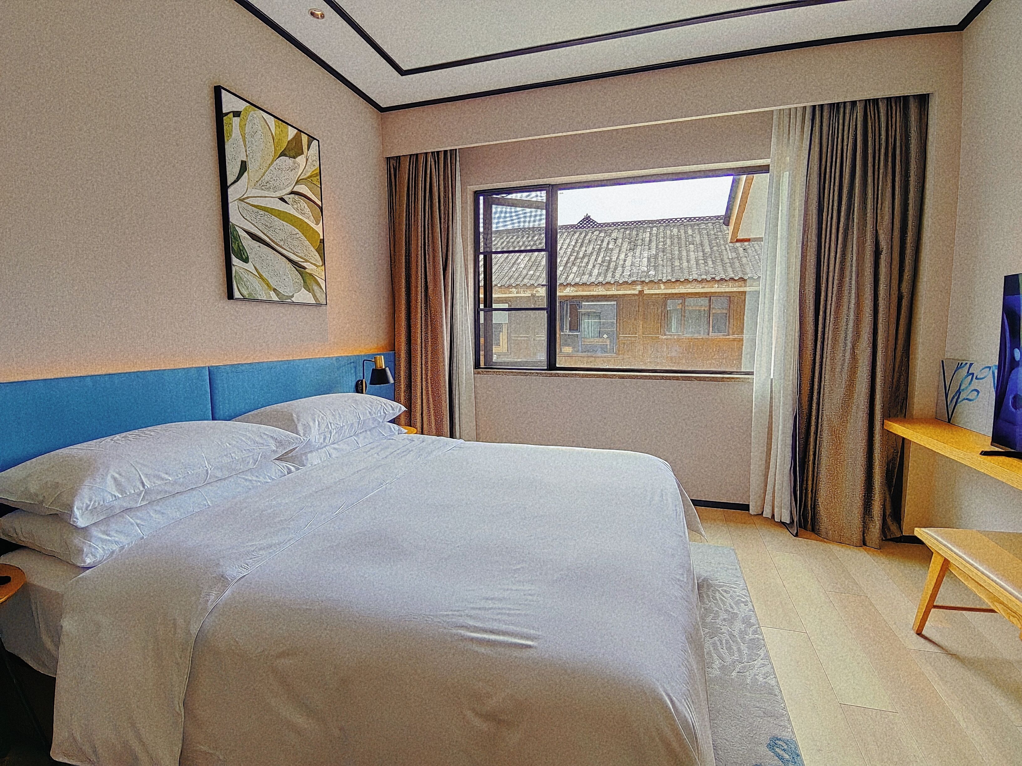 A hotel room with a large bed, a bedside table, a window with curtains, and a view of buildings outside.