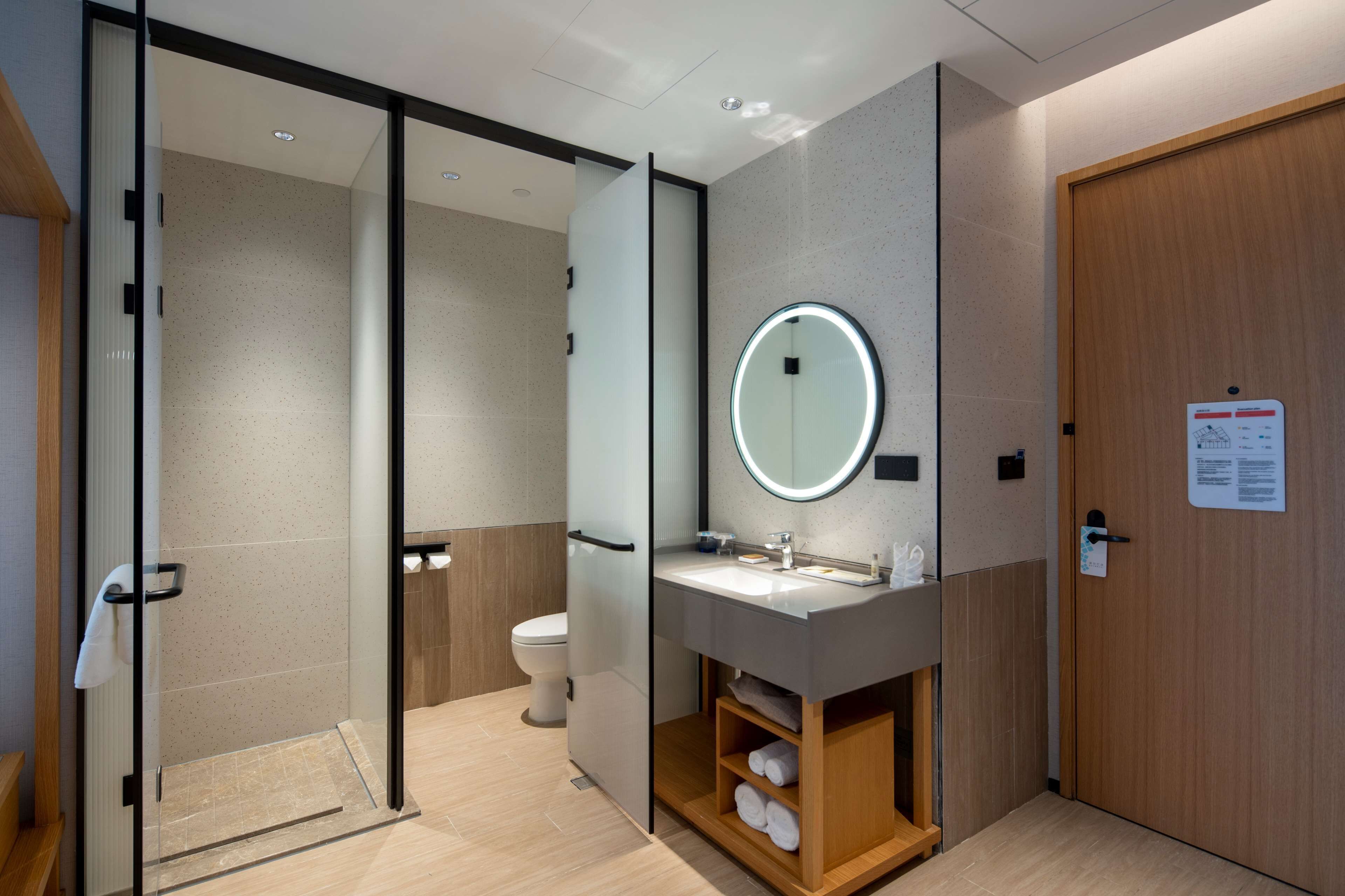 Un baño moderno con cabina de ducha de vidrio, inodoro, lavabo con espejo y estante de madera con toallas.