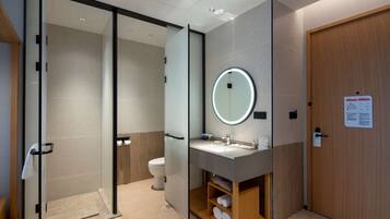 Un baño moderno con cabina de ducha de vidrio, inodoro, lavabo con espejo y estante de madera con toallas.
