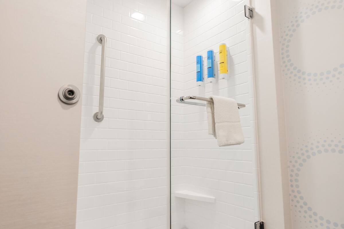 Een douche met glazen wanden, een betegelde muur in het wit, een handdoekrek met een witte handdoek en twee flessen aan de muur.