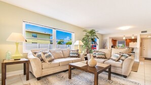 A living room with a beige sofa set, two wooden side tables, a patterned rug, and a view of a building through large windows. - Maximo Marina Condo • Heated pool, Near Pass-a-Grille, Downtown & Eckerd (St. Petersburg)