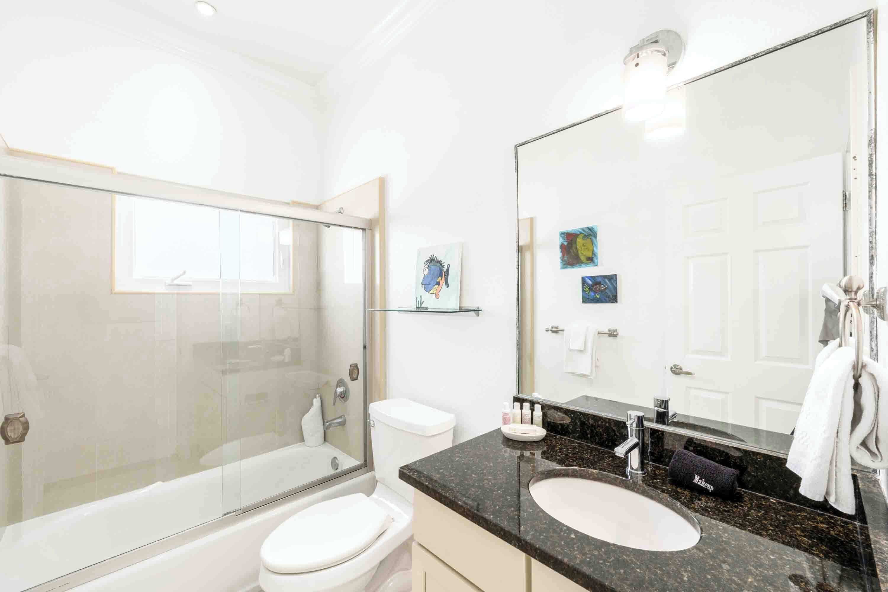 Une salle de bain moderne avec une douche à parois de verre, des toilettes blanches, un lavabo avec un grand miroir et un meuble-lavabo avec des articles de toilette.