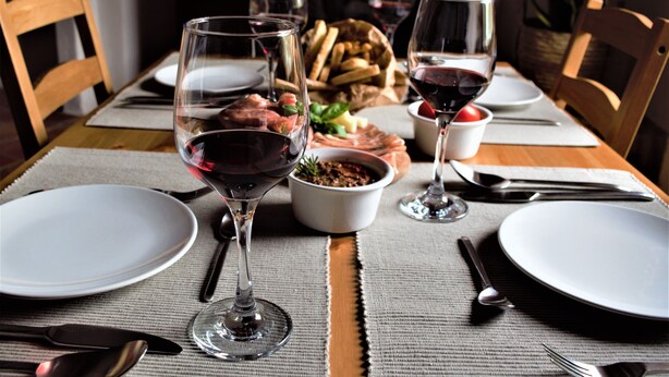 A dining table set with plates, cutlery, and wine glasses, with a bowl of bread and a dish of food in the center.