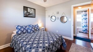 Quarto com cama, ventoinha no teto, mesa de cabeceira, espelho fixado na parede e porta para um banheiro.