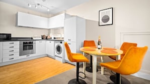 Een moderne keuken met witte kastjes, een magnetron, oven en wasmachine. Er is een eethoek met een tafel, stoelen en een fles champagne.