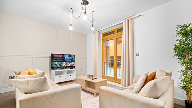 A modern living room with a flat-screen TV, two beige sofas, a coffee table, and a large window with curtains.