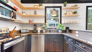 A modern kitchen with dark cabinets, a built-in oven, a dishwasher, and a granite countertop. There are wooden shelves above the sink and a window above the sink.