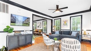 A modern living room with a grey sofa, a white armchair, and a coffee table. There is a television mounted on the wall, a ceiling fan, and a cabinet with decorative items.