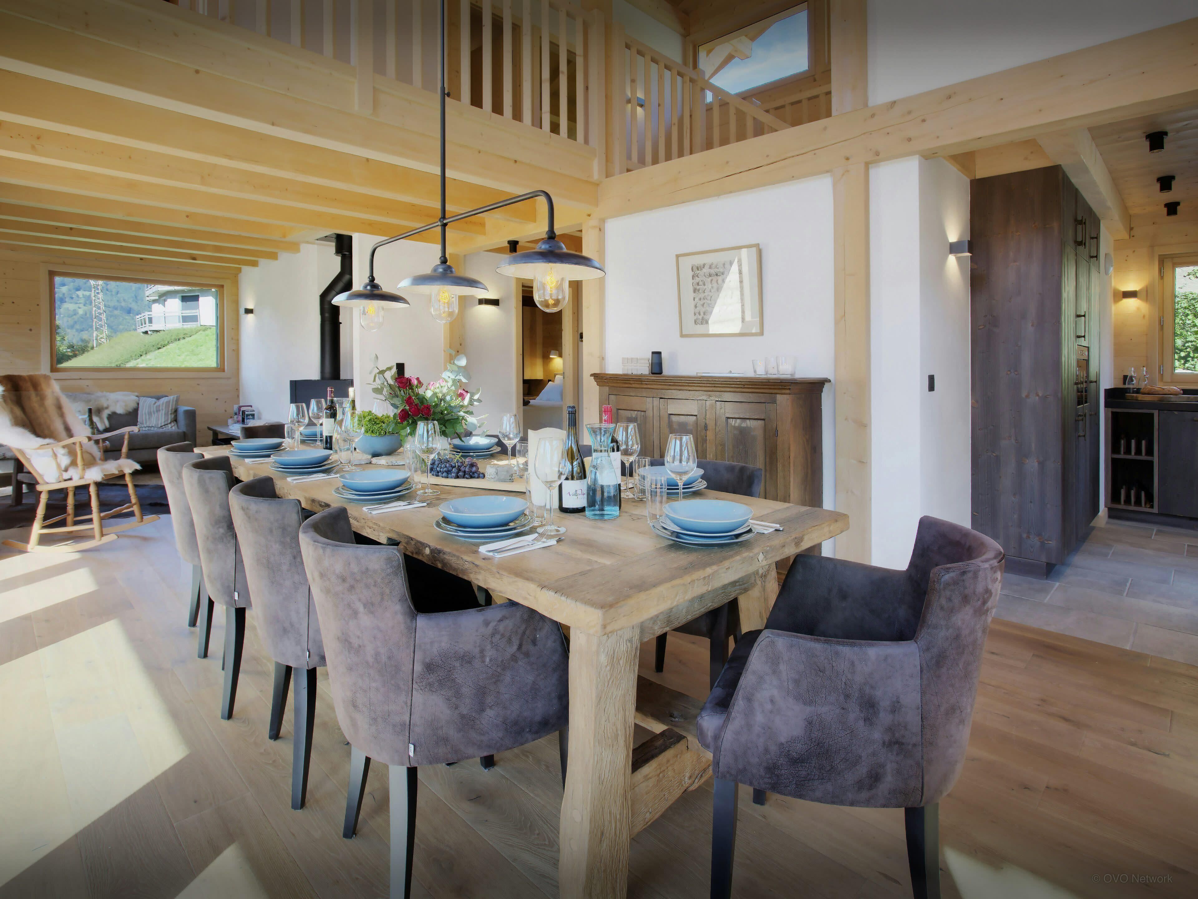 A dining area with a wooden table set for four, surrounded by chairs, and a view of the outdoors through a large window.