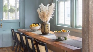 Un coin repas comprenant une table en bois, quatre chaises, un vase contenant des plantes séchées et un saladier rempli de fruits.