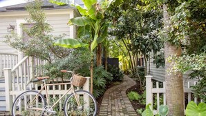 Un vélo est garé sur un chemin pavé de briques, bordé d’une clôture en bois peinte en blanc et entouré d’une végétation luxuriante.