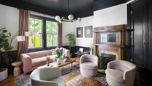 A living room with a black ceiling, a fireplace, a pink sofa, and a large window with curtains. - The Monroe | Award-Winning Renovated Historic Home (Charleston)