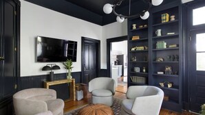 A living room with a black and white color scheme, featuring a large TV, multiple armchairs, a coffee table, and a bookshelf. - The Monroe | Award-Winning Renovated Historic Home (Charleston)