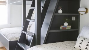 A bunk bed room with a ladder, a shelf with books and a vase, and a bedside table with a lamp. - The Monroe | Award-Winning Renovated Historic Home (Charleston)