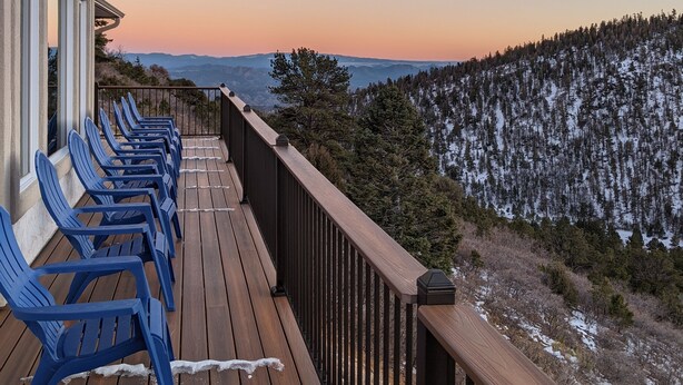 A wooden deck with blue chairs, overlooking a snowy forest and distant mountains during sunset.