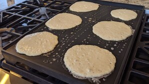 A kitchen with a griddle cooking pancakes, a vase with flowers, and a stone wall.