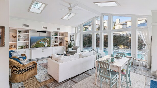 A spacious living area with a white sofa, dining table, and chairs, a television, and a view of a swimming pool and greenery. - Big East Beach Home with Pool (Saint Simons Island)