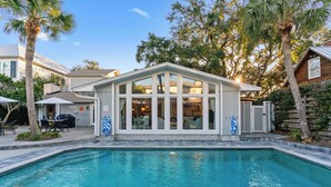 A swimming pool with a covered patio area, surrounded by palm trees and a wooden cabin. - Big East Beach Home with Pool (Saint Simons Island)