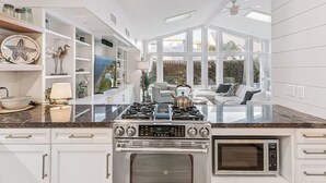 A modern kitchen with white cabinetry, a gas stove, an oven, and a microwave. There is a large window providing a view of the outdoors. - Big East Beach Home with Pool (Saint Simons Island)