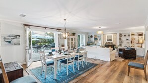 A spacious living area with a dining table, chairs, and a sofa. There is a kitchen area with cabinets and a countertop. The room has a large window and a view of the outdoors. - Big East Beach Home with Pool (Saint Simons Island)