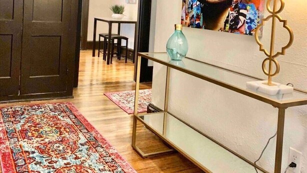 A room with a dark wooden wardrobe, a colorful abstract painting on the wall, a glass console table with a lamp, and a patterned rug on the floor. - Modern Midtown Retreat (Montgomery)