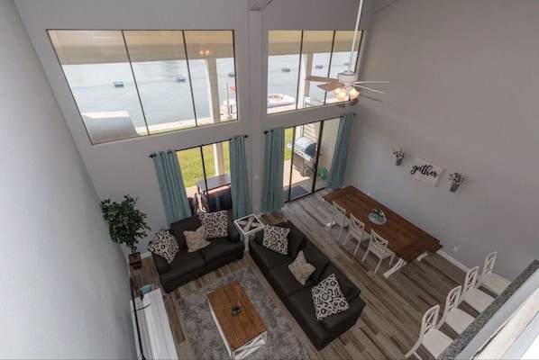 A spacious living area with a large window overlooking a body of water, a dining area with a table and chairs, and a ceiling fan.