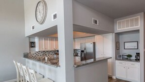 A modern kitchen with white cabinets, a stainless steel refrigerator, and a granite countertop. There are bar stools and a clock on the wall.