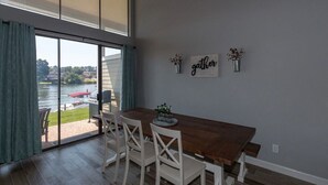 A dining area with a wooden table and white chairs, large windows with curtains, and a view of a lake with a boat.