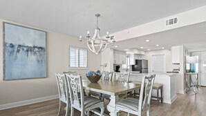 A dining area with a large table, chairs, and a chandelier. There is a kitchen in the background with cabinets and appliances. - Seas the day - Destin Gulfgate 1006! (Destin)