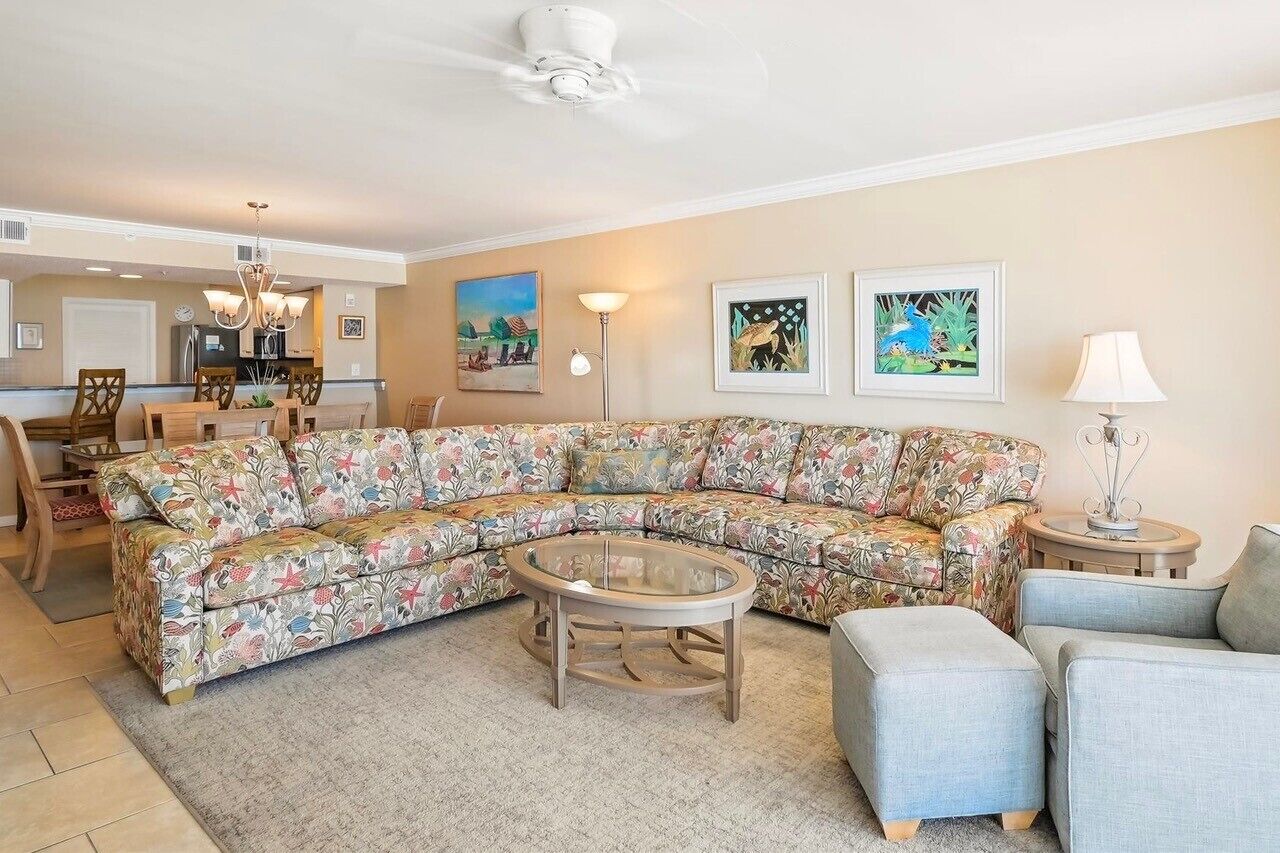 A living room with a floral sofa, a glass coffee table, and a grey armchair. There is a dining area in the background with a chandelier.