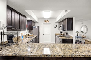 A kitchen with granite countertops, dark wooden cabinets, a white door, and a clock on the wall.
